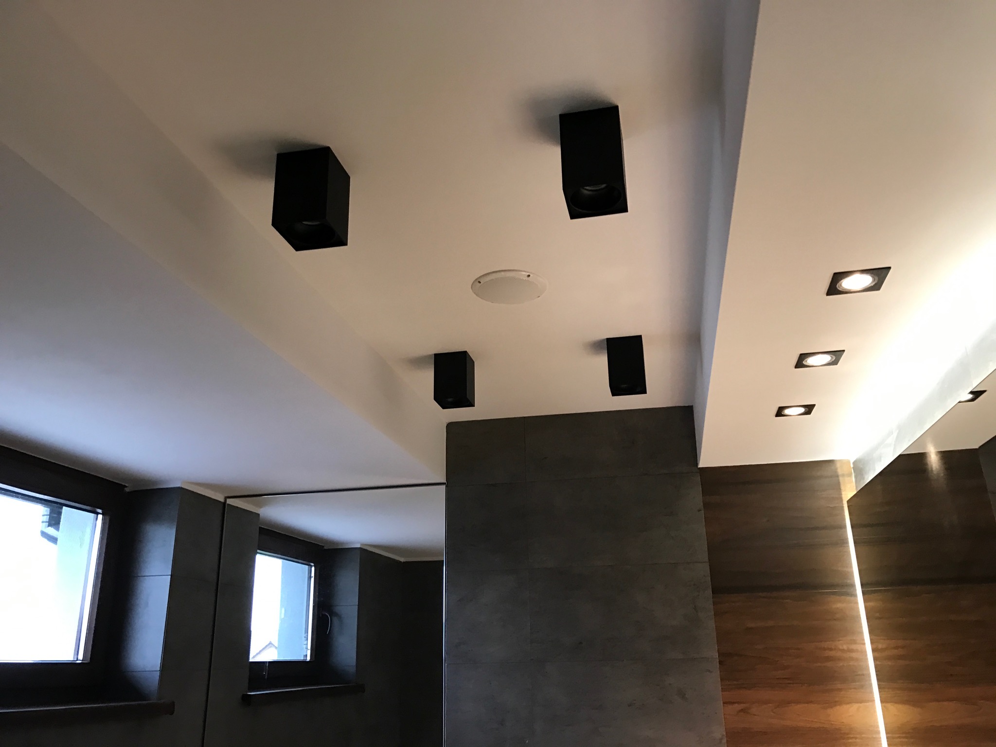 Nowoczesna łazienka z czarnymi, kubicznymi lampami sufitowymi i podłużnymi panelami z oświetleniem LED, szare płytki na ścianie i okno.