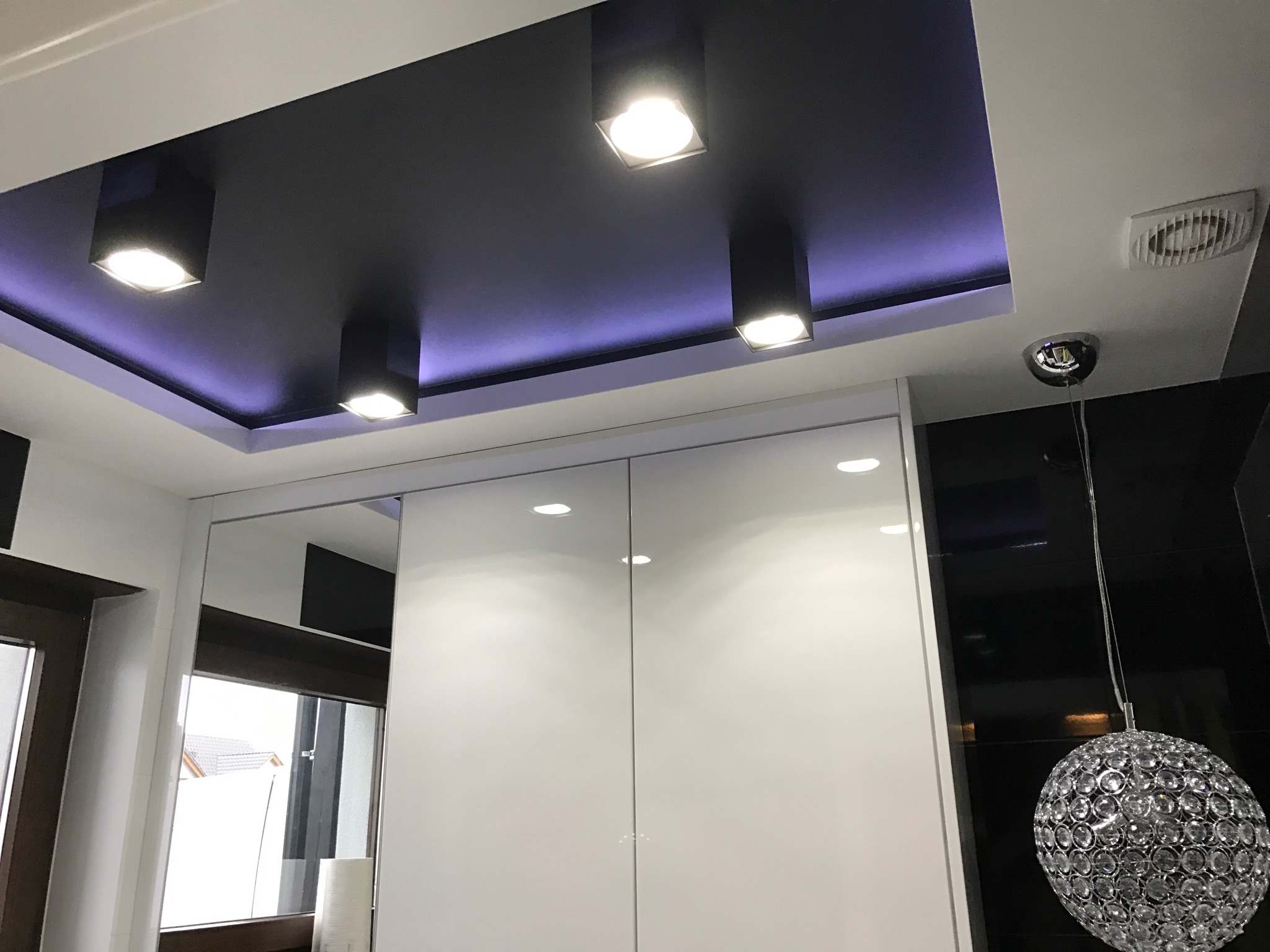 Nowoczesne oświetlenie sufitowe z czarną wnęką i listwą LED, biała zabudowa meblowa z przesuwnymi drzwiami, oraz dekoracyjna lampa wisząca w kształcie kuli.