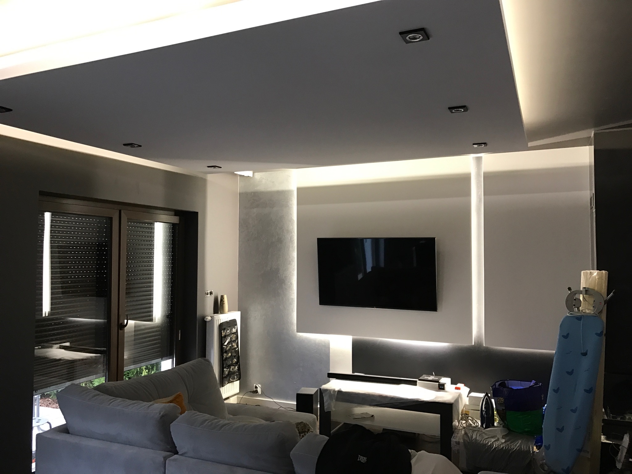 Nowoczesny salon z szarą sofą, oświetleniem LED na suficie i ścianie za telewizorem, oraz zasłoniętym oknem.