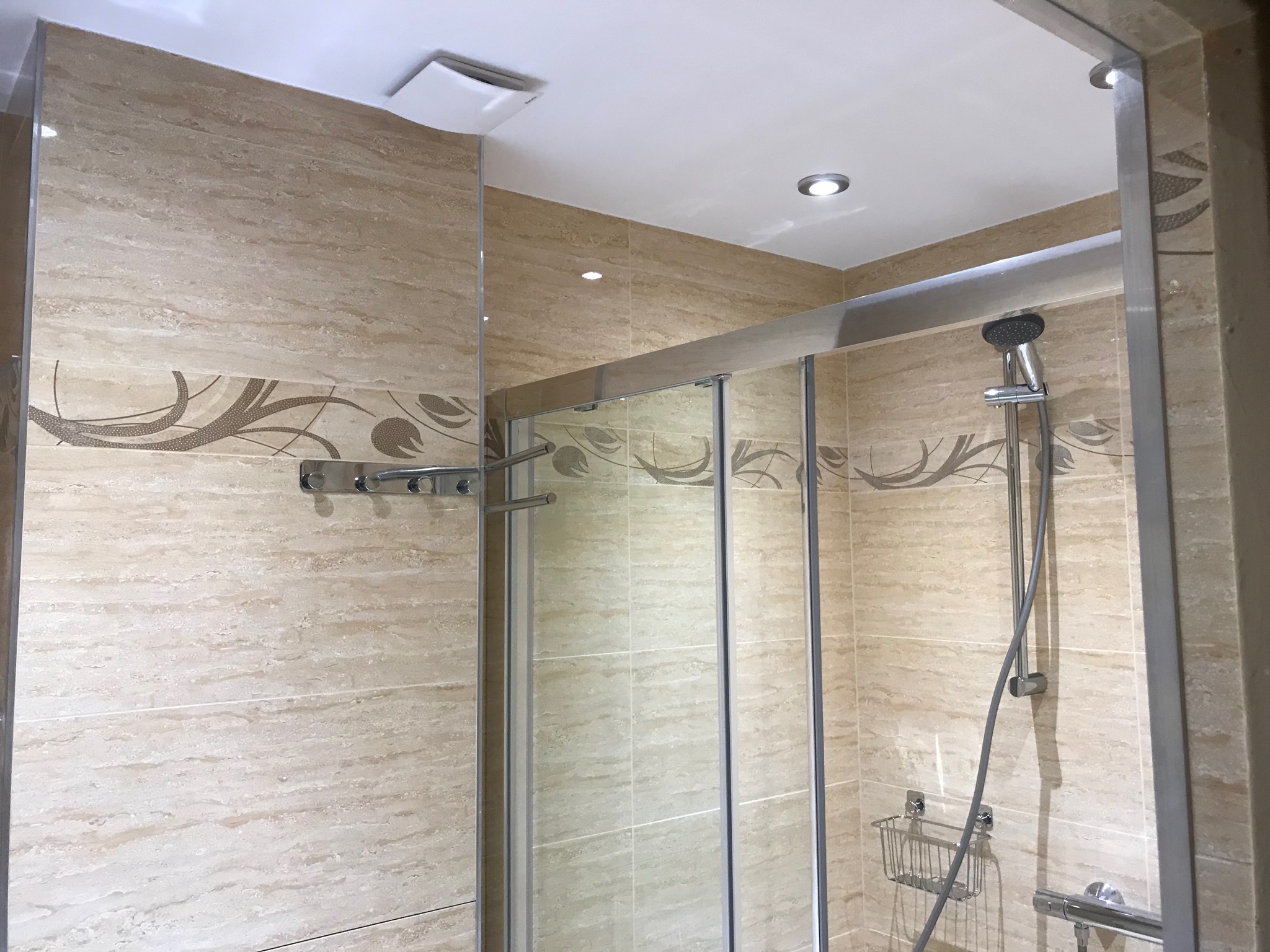 Beżowa łazienka z kabiną prysznicową, wykończona płytkami imitującymi trawertyn z delikatnym, roślinnym ornamentem, oświetleniem punktowym i chromowanymi akcesoriami.