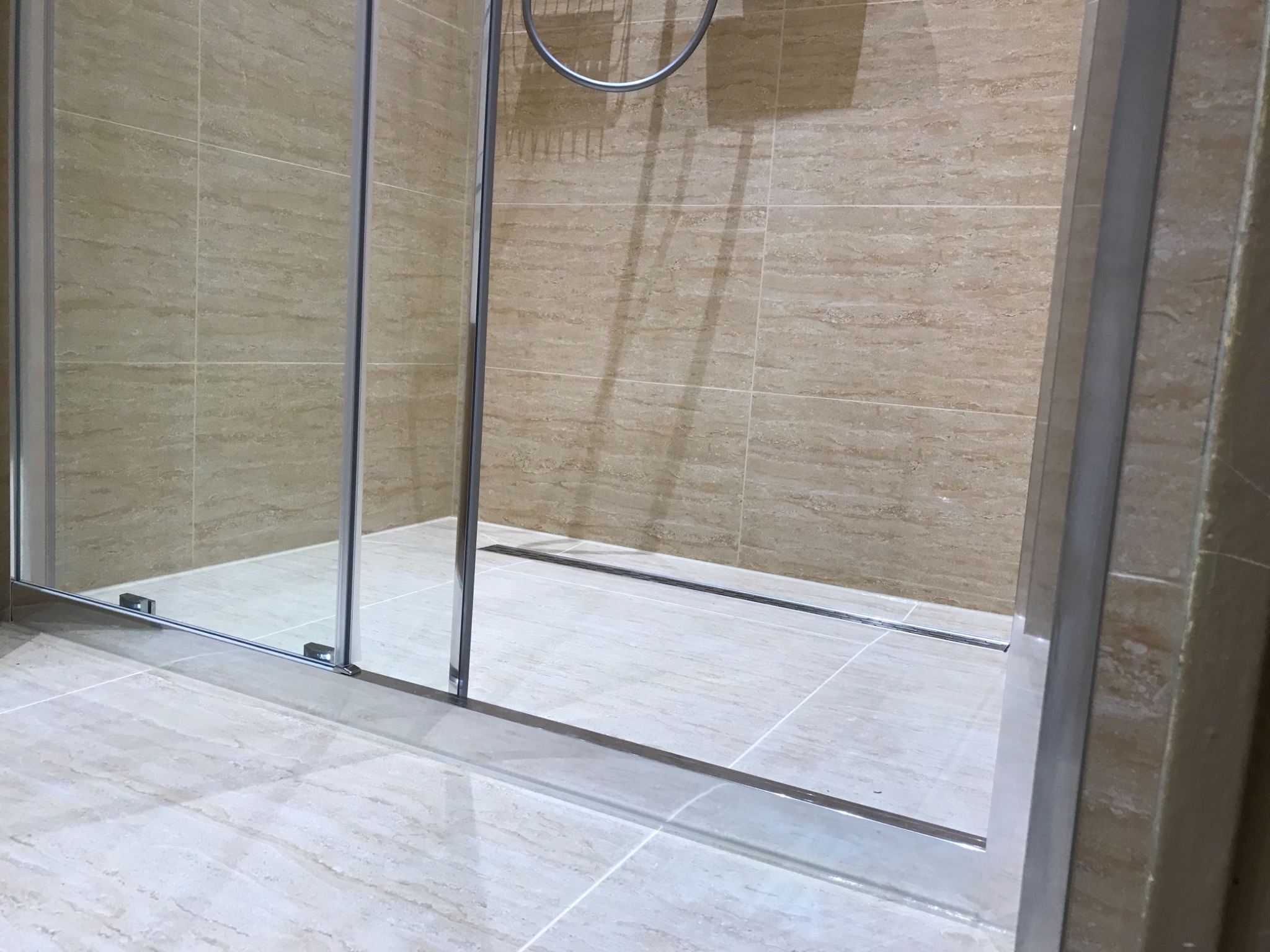 Beżowa łazienka z kabiną prysznicową typu walk-in, widoczne płytki imitujące trawertyn na ścianach i podłodze, oraz odpływ liniowy.