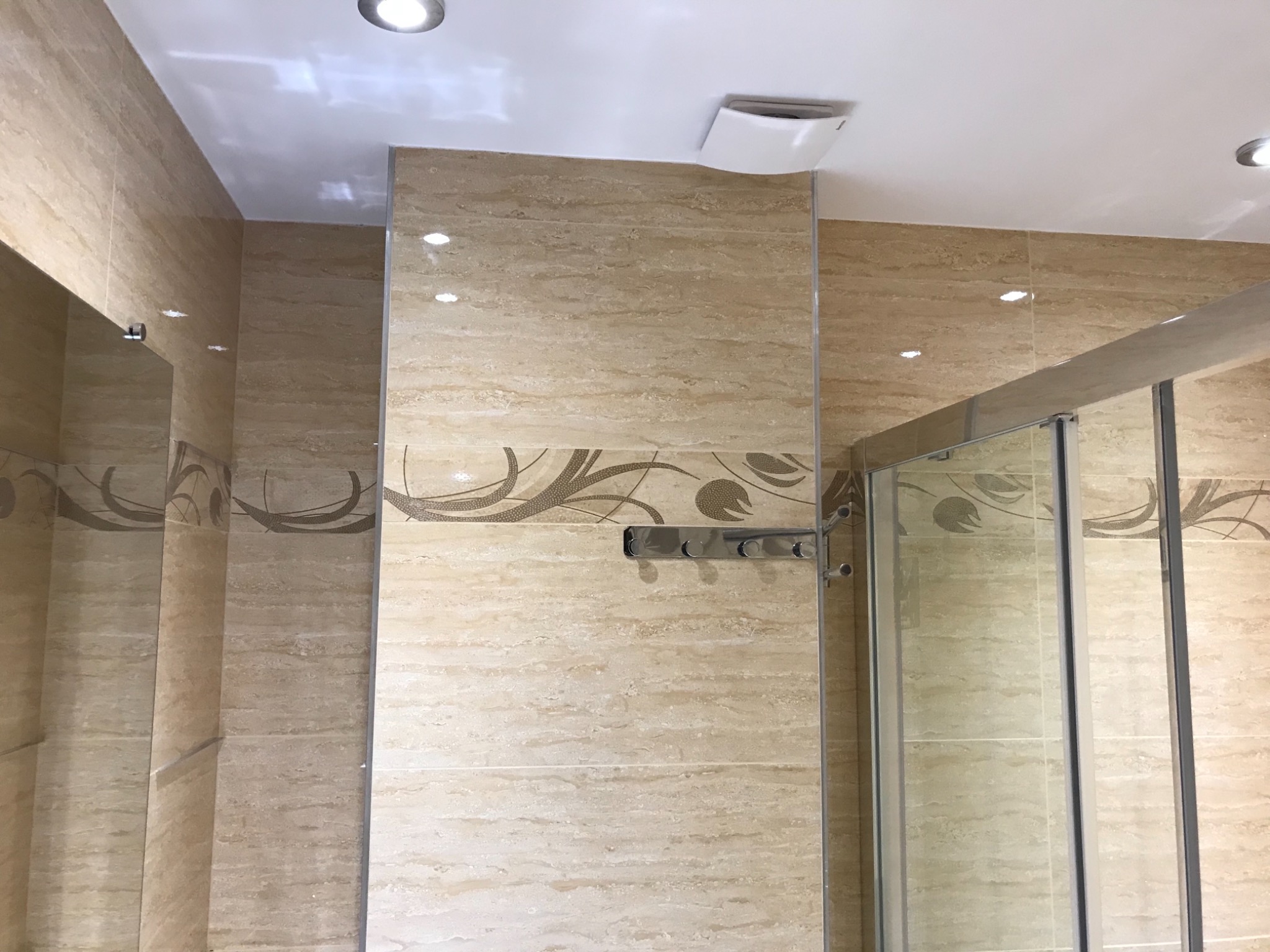 Wykończona łazienka z beżowymi, marmurkowymi płytkami z dekoracyjnym pasem, kabina prysznicowa ze szklanymi drzwiami, oświetlenie punktowe w suficie i metalowy wieszak na ręczniki.