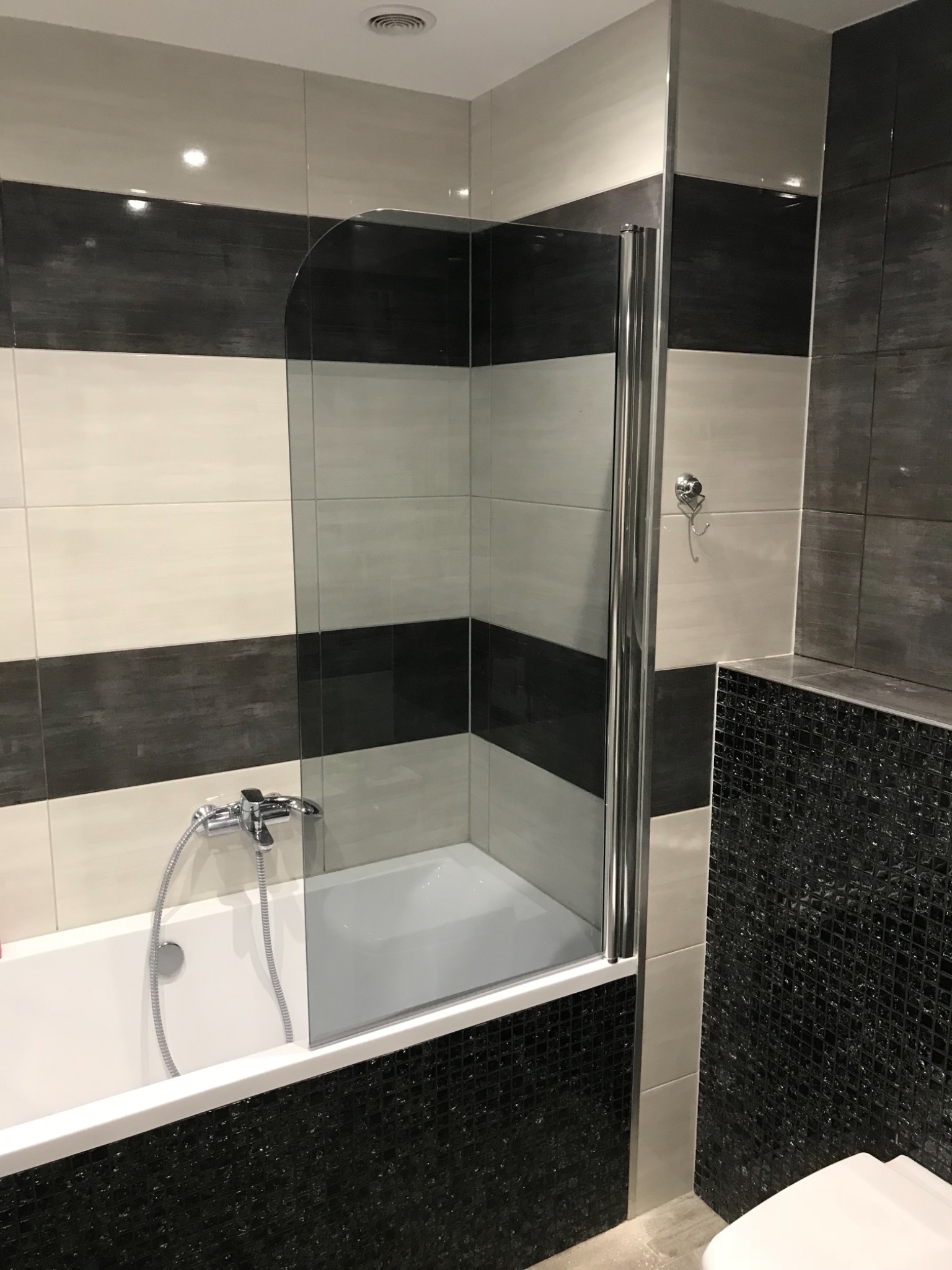 Nowoczesna łazienka we Wrocławiu z wanną obudowaną czarno-białą mozaiką, szklanym parawanem nawannowym z chromowanymi profilami oraz ścianami wyłożonymi pasami płytek w odcieniach szarości i bieli.