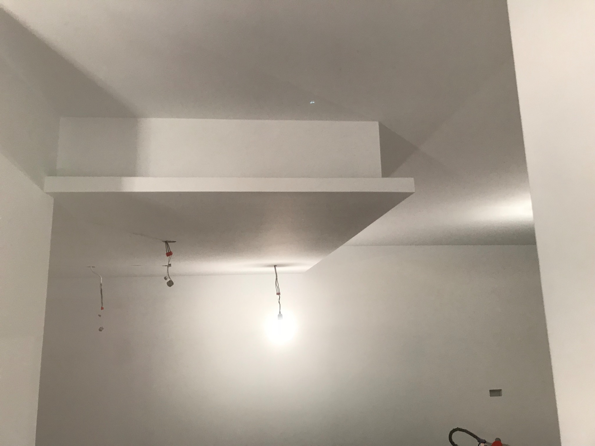 Wnętrze w trakcie wykończenia: białe ściany, podwieszony sufit z wystającym elementem, wiszące kable oświetleniowe, widoczne gniazdko elektryczne, narzędzie na podłodze.
