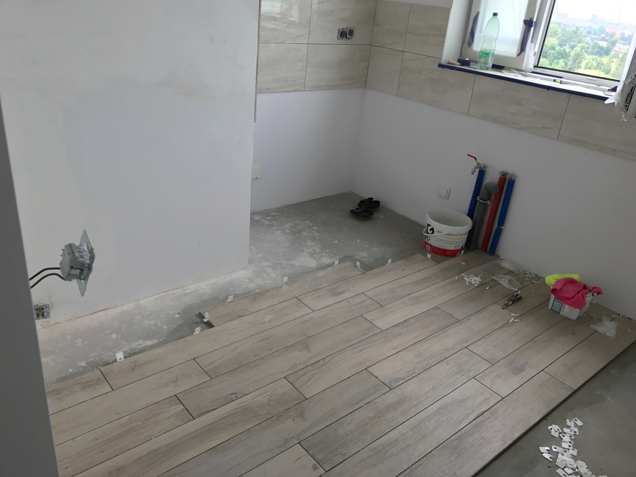 Układanie podłogi z płytek imitujących drewno w łazience, widoczne krzyżyki dystansowe, wiadro z zaprawą i narzędzia glazurnicze.
