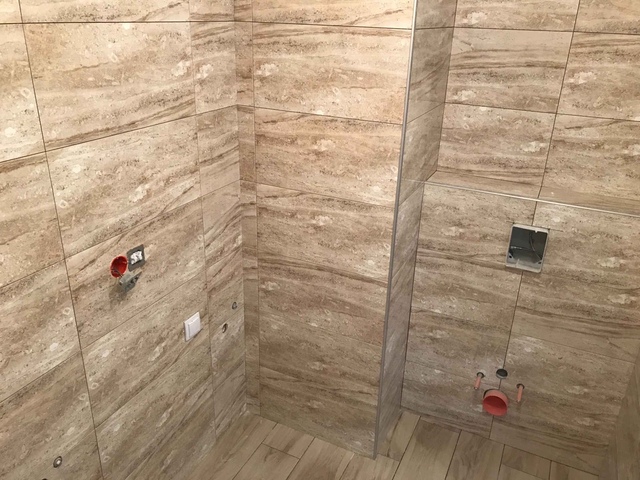 Łazienka w trakcie remontu: beżowe płytki imitujące trawertyn na ścianach i podłodze, widoczne instalacje elektryczne i hydrauliczne przed montażem armatury.