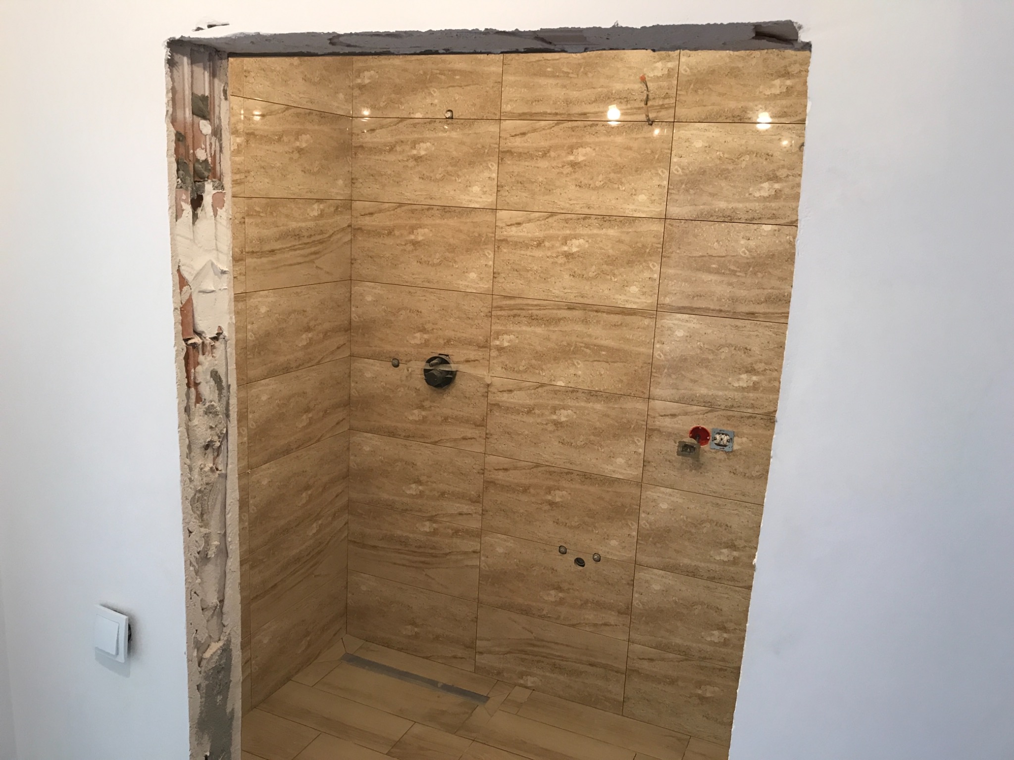 Wnętrze łazienki w trakcie remontu, widoczne beżowe płytki imitujące marmur na ścianach i podłodze, surowy otwór drzwiowy z odsłoniętym murem i resztkami zaprawy, instalacja wodna i elektryczna...