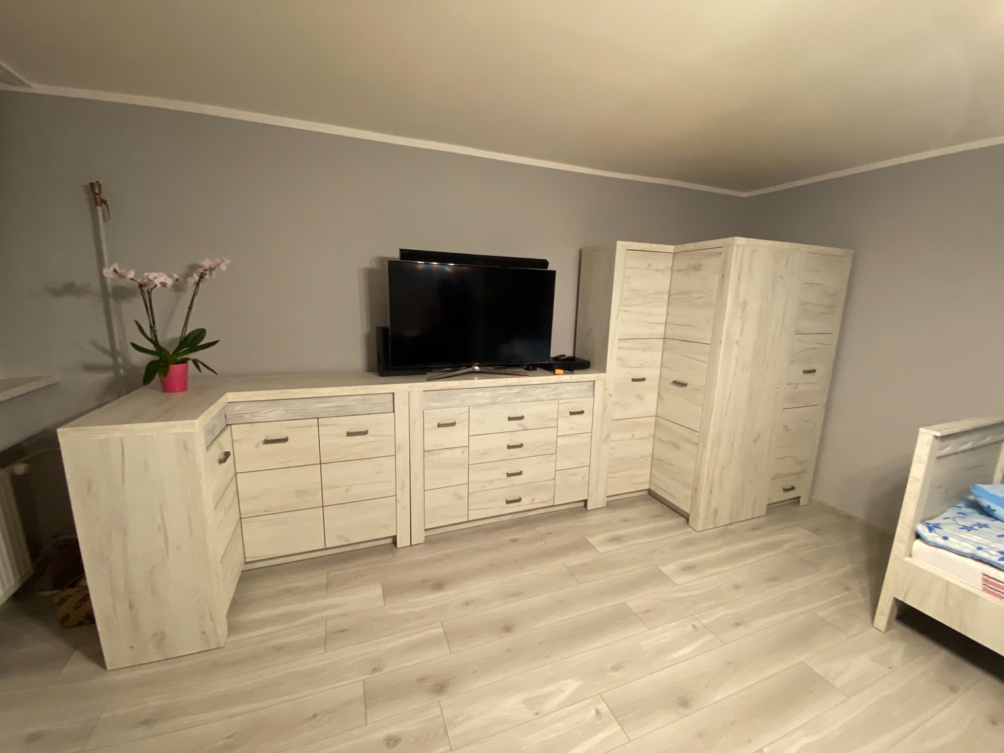 Jasne meble z drewnopodobnym wzorem, w tym szafki, komody i wysoka szafa, ustawione na podłodze z paneli, z telewizorem na komodzie i kwiatami w doniczce na blacie.