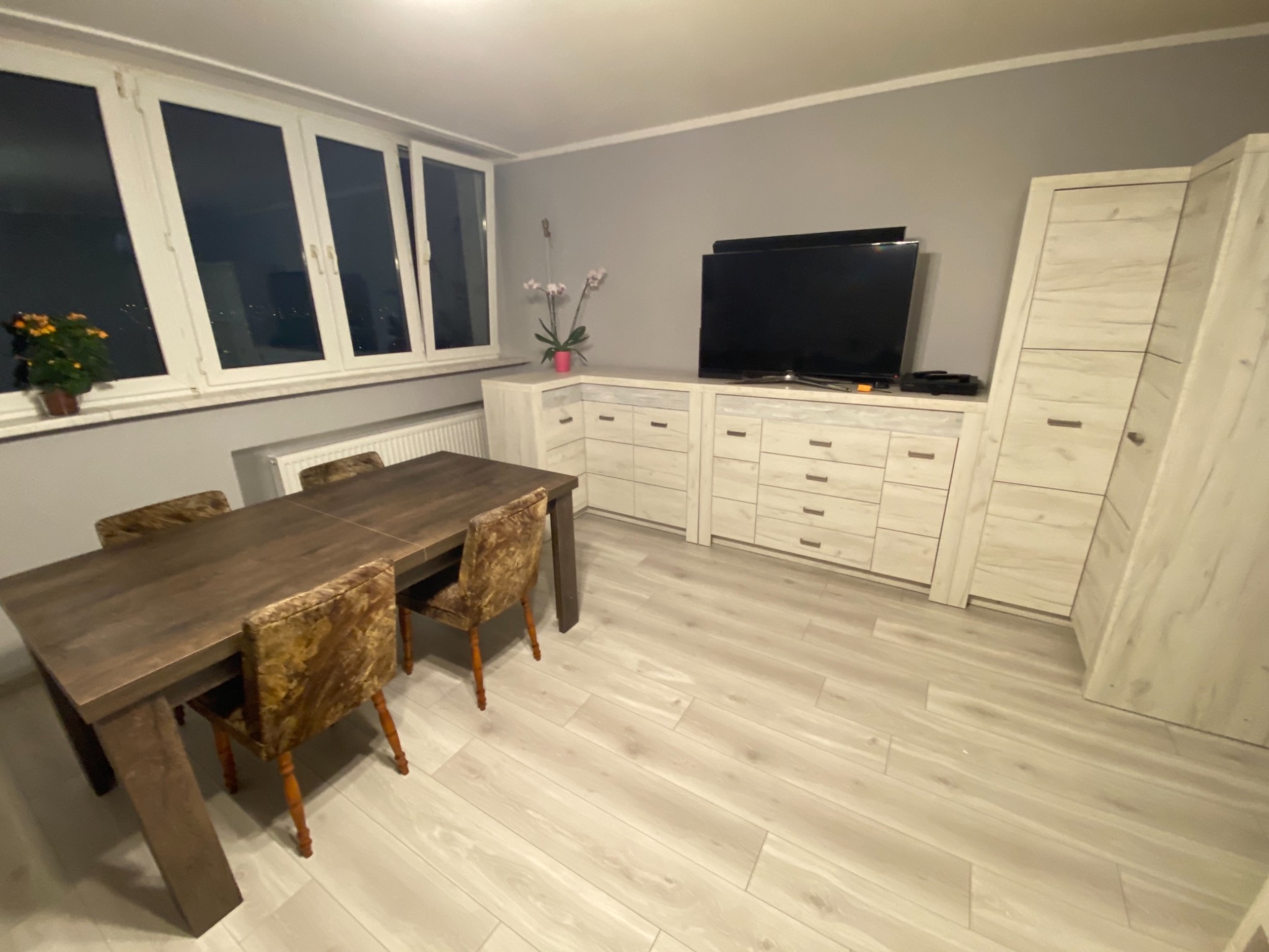 Jadalnia z drewnianym stołem i krzesłami obitymi wzorzystym materiałem, na podłodze panele imitujące drewno, w tle białe meble z szufladami i szafka.