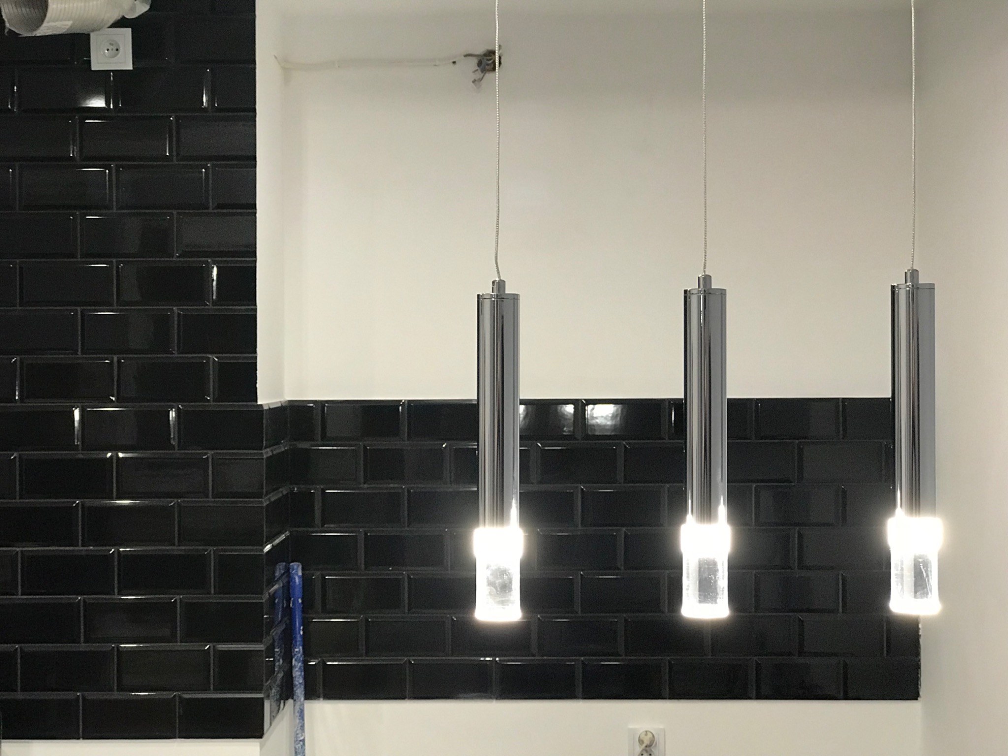 Błyszczące, czarne płytki ułożone w cegiełkę na ścianie, oświetlone przez trzy nowoczesne, chromowane lampy wiszące z białym światłem.