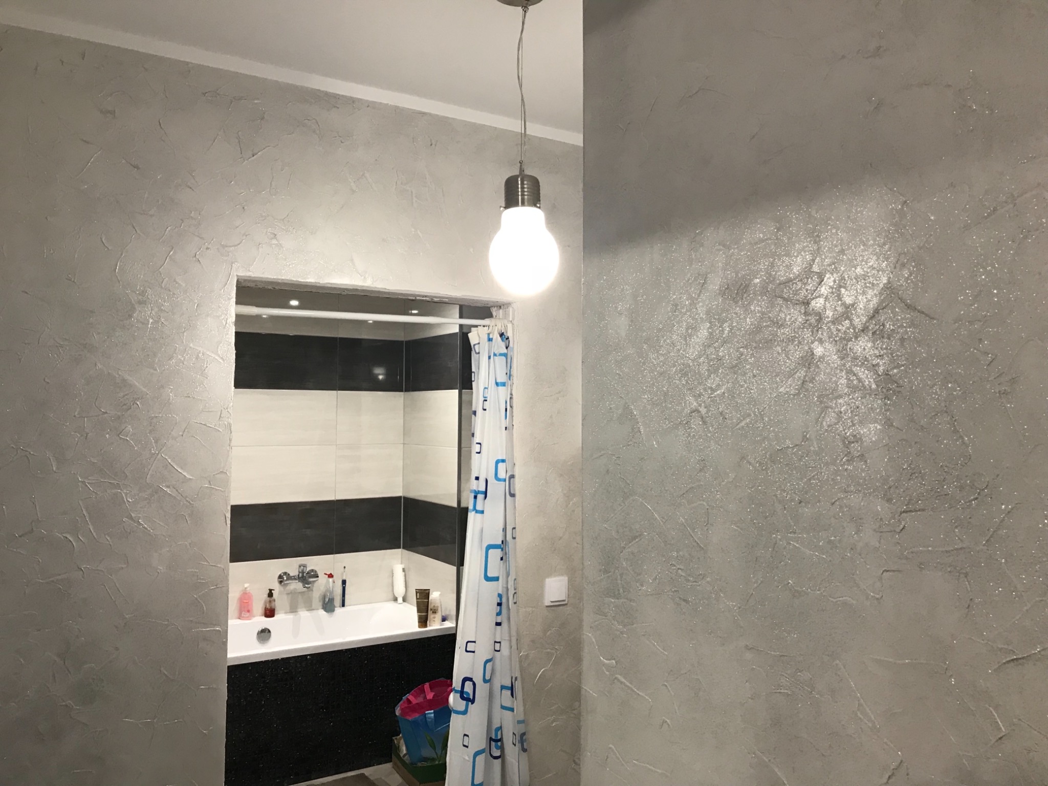 Ściana w łazience z dekoracyjnym szpachlowaniem w odcieniach szarości, widoczna wanna z czarno-białymi płytkami i zasłona prysznicowa w niebieskie wzory, oświetlona wiszącą lampą w kształcie żarówki.