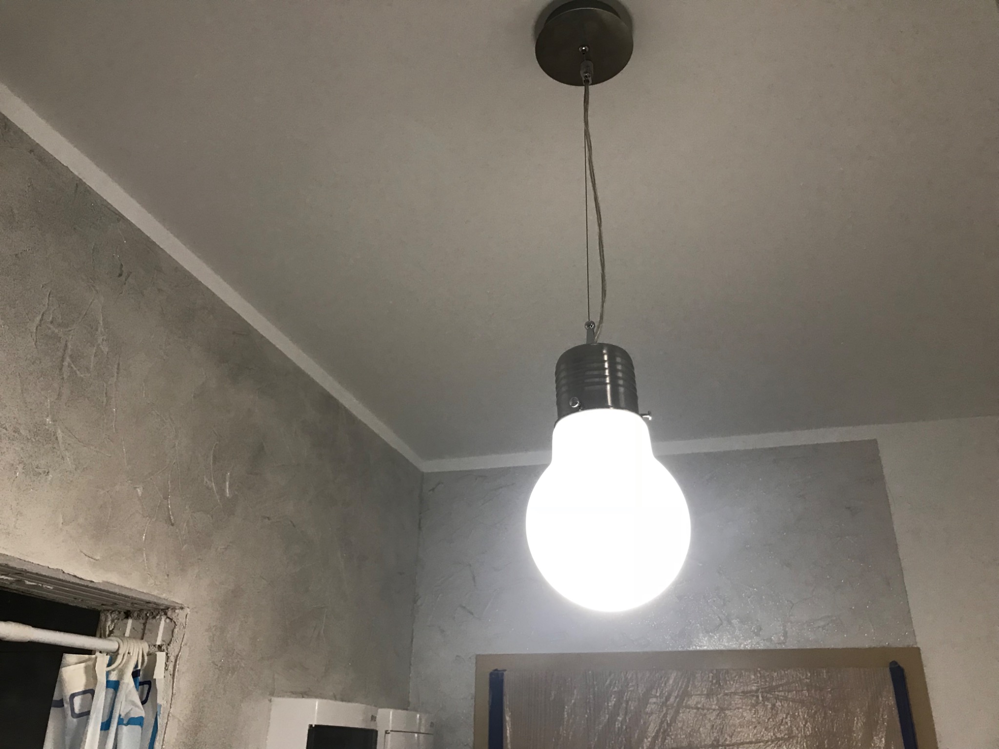 Lampa wisząca w kształcie żarówki oświetla szarą ścianę z efektem dekoracyjnym, widoczny fragment sufitu z listwą oraz zabezpieczone folią drzwi.