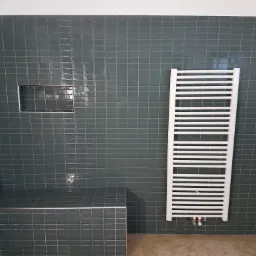 Łazienka wykończona ciemnymi, kwadratowymi płytkami, z wnęką w ścianie i białym grzejnikiem drabinkowym. Widoczna także zabudowana półka wyłożona tymi samymi płytkami.