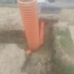 Pomarańczowa karbowana rura pionowa połączona z poziomą rurą w wykopie ziemnym, widoczne fragmenty geowłókniny i trawy.