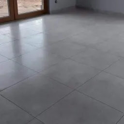 Wnętrze pokoju z nową, szarą podłogą wyłożoną kwadratowymi płytkami, widoczne przesuwne drzwi balkonowe z brązowymi ramami i szare ściany z gładzią, w tle widok na ogród.