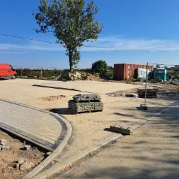 Nowa nawierzchnia z kostki brukowej w trakcie budowy, paleta z kostką na środku placu, widoczny czerwony van i kontener w tle, teren budowy w słoneczny dzień.