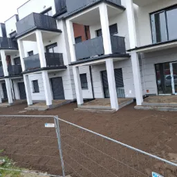 Nowe apartamenty z balkonami i przygotowanym terenem pod trawnik, widoczne ślady po pracach ziemnych oraz tymczasowe ogrodzenie zabezpieczające.