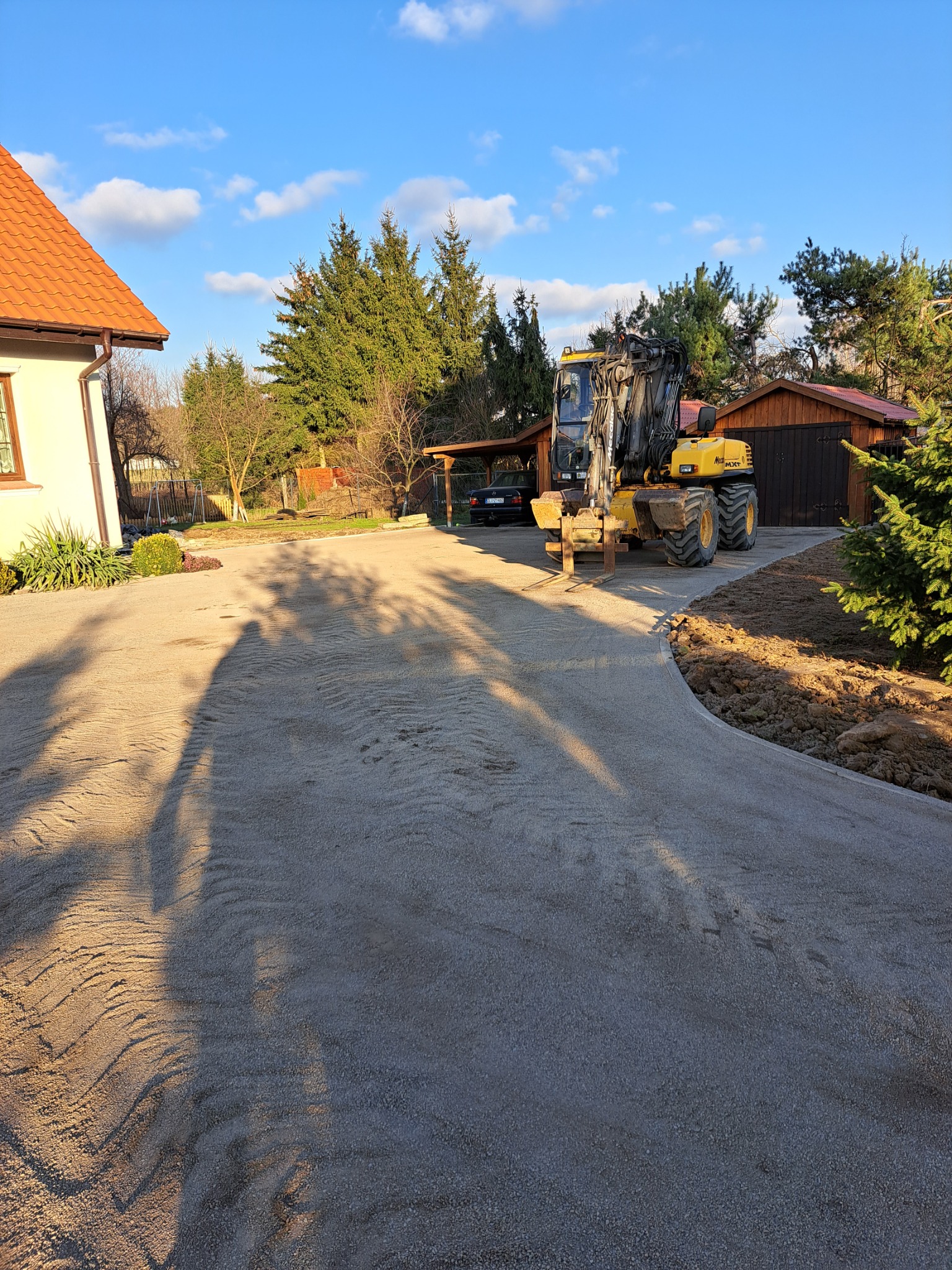 Żółta koparka kołowa na utwardzonej drodze dojazdowej, przygotowanej pod kostkę brukową, widoczny dom i garaż w tle.