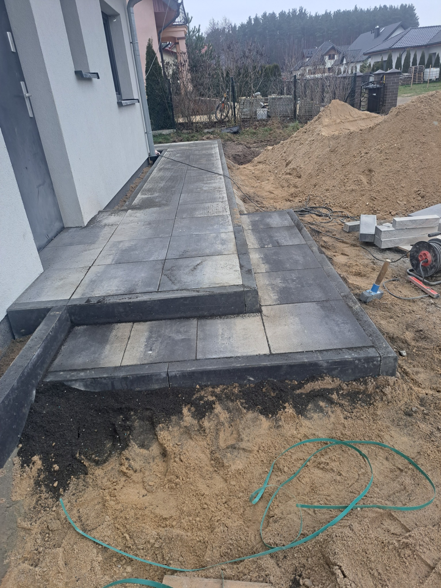 Ułożona ścieżka z szarych płyt betonowych prowadząca do drzwi budynku, z dwoma stopniami. W tle hałda piasku i narzędzia budowlane.