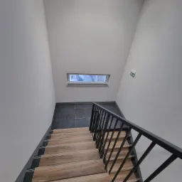 Wnętrze klatki schodowej z drewnopodobnymi stopniami, ciemnymi płytkami na podłodze i czarną metalową balustradą, widok z góry, okno dachowe i znak ewakuacyjny na ścianie.