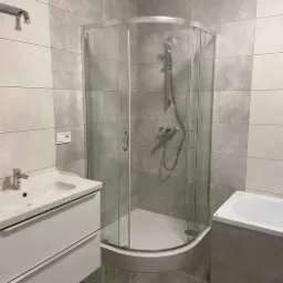 Nowoczesna łazienka z kabiną prysznicową narożną, umywalką z szafką w kolorze białym i wanną obudowaną szarymi płytkami, wykończona szarą płytką imitującą kamień.