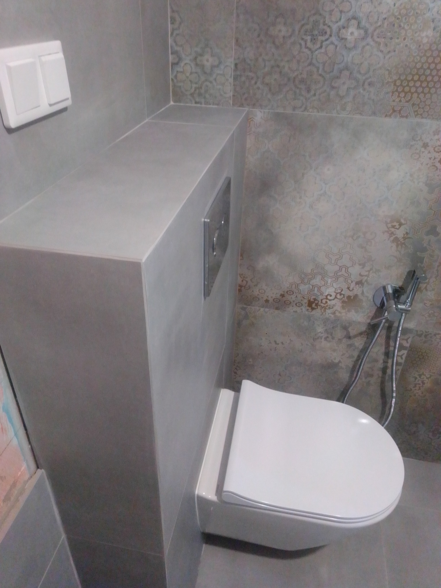 Podwieszana toaleta z zabudową w szarej tonacji, ozdobne płytki na ścianie, bidetka w komplecie.