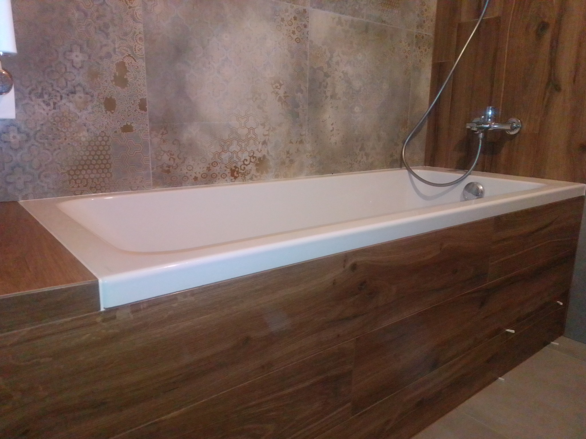 Wykończona obudowa wanny z paneli drewnopodobnych, bateria natryskowa na ścianie z wzorzystych płytek, widoczne fugi i listwy wykończeniowe.