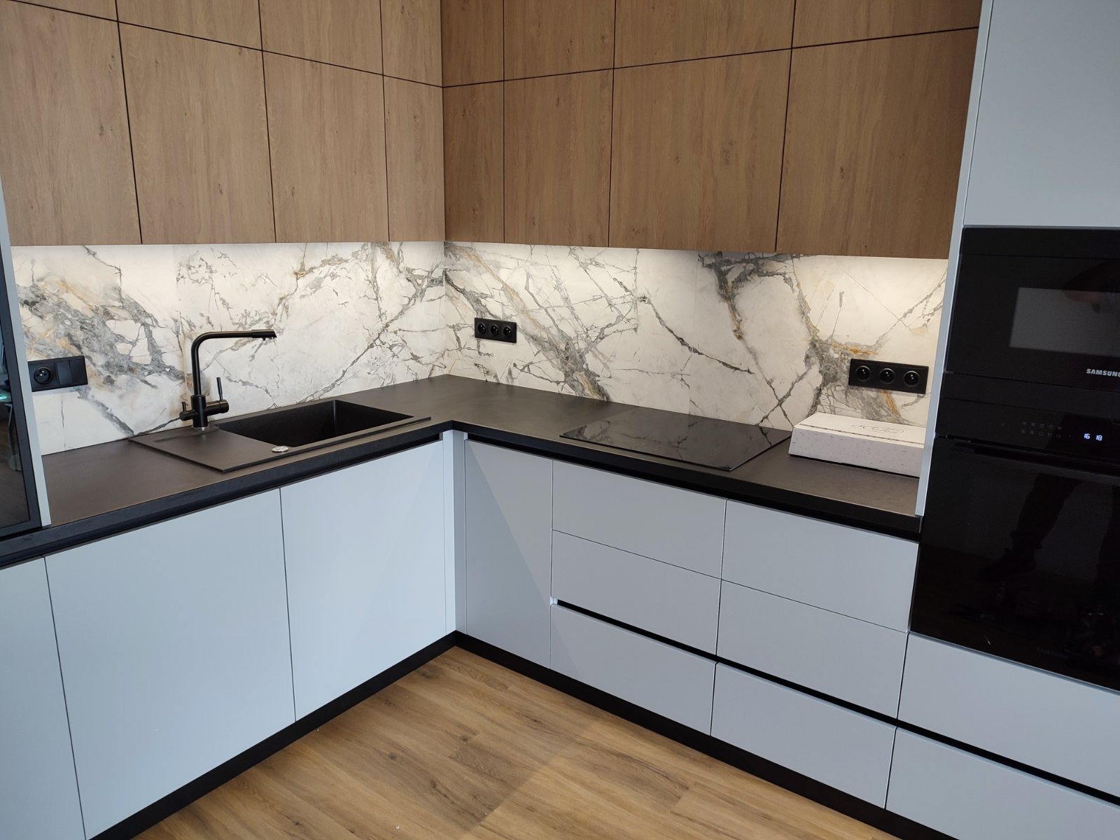 Nowoczesna kuchnia z białymi szafkami, czarnym blatem i marmurowym panelem ściennym, widoczny zlew z czarną armaturą i płyta indukcyjna.