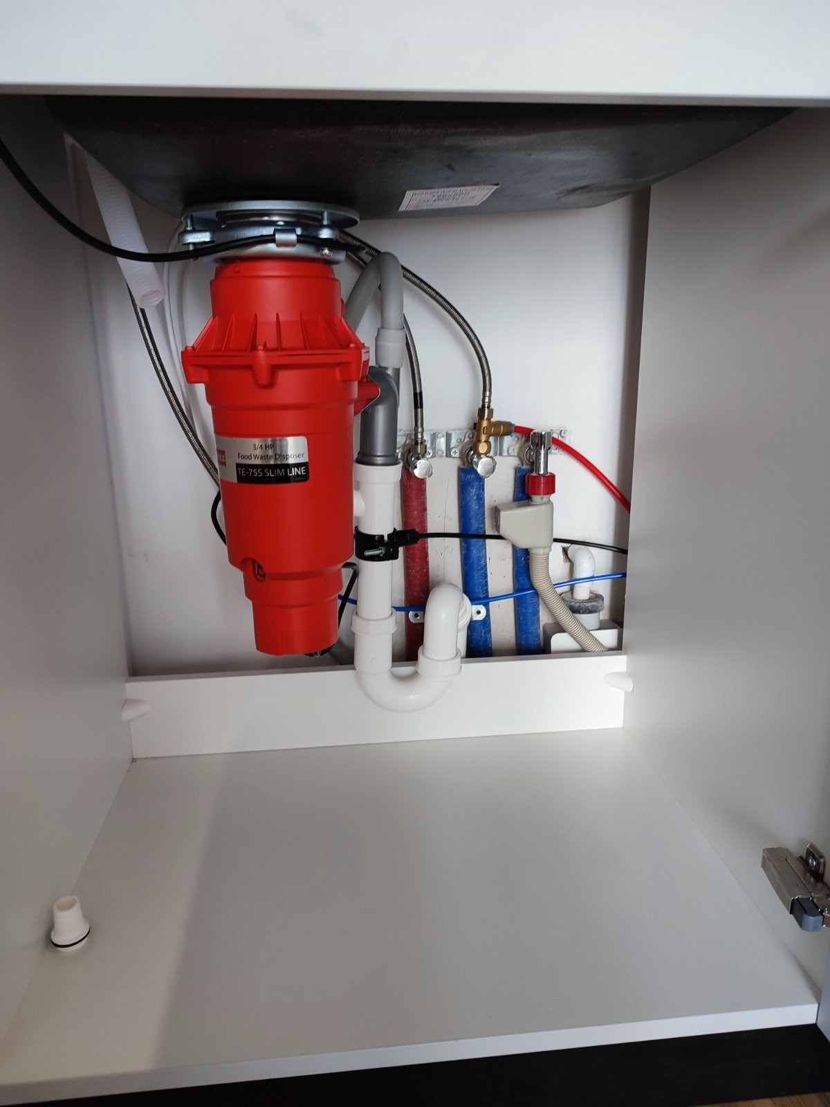 Czerwony rozdrabniacz do odpadów zamontowany pod zlewem w szafce, widoczne rury doprowadzające i odprowadzające wodę.
