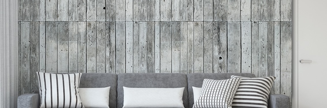 Szary narożnik z poduszkami na tle ściany z desek o efekcie postarzenia, z widoczną białą ścianą i drzwiami po prawej stronie.