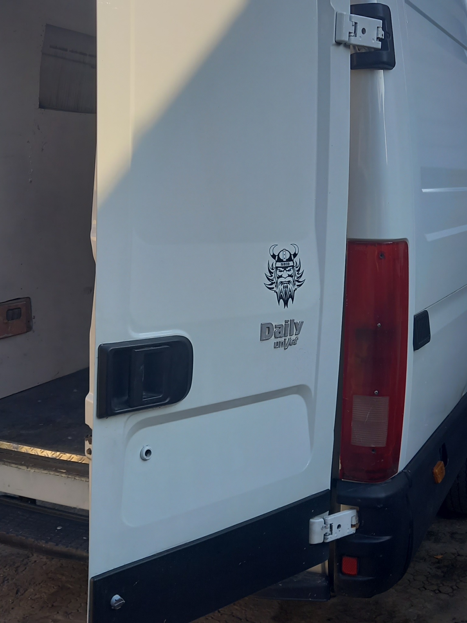 Otwarta tylna klapa białego busa marki Iveco Daily z naklejką w kształcie wikinga, zaparkowanego w Radomiu, widok z tyłu, ukazujący częściowo wnętrze przestrzeni ładunkowej.
