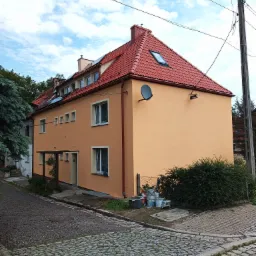 Dwupiętrowy budynek z pomarańczową elewacją i czerwoną dach&oacute;wką, widoczny z poziomu brukowanej ulicy. W tle błękitne niebo z chmurami. Przy ścianie budynku widoczne wiaderka z farbą i krzew.