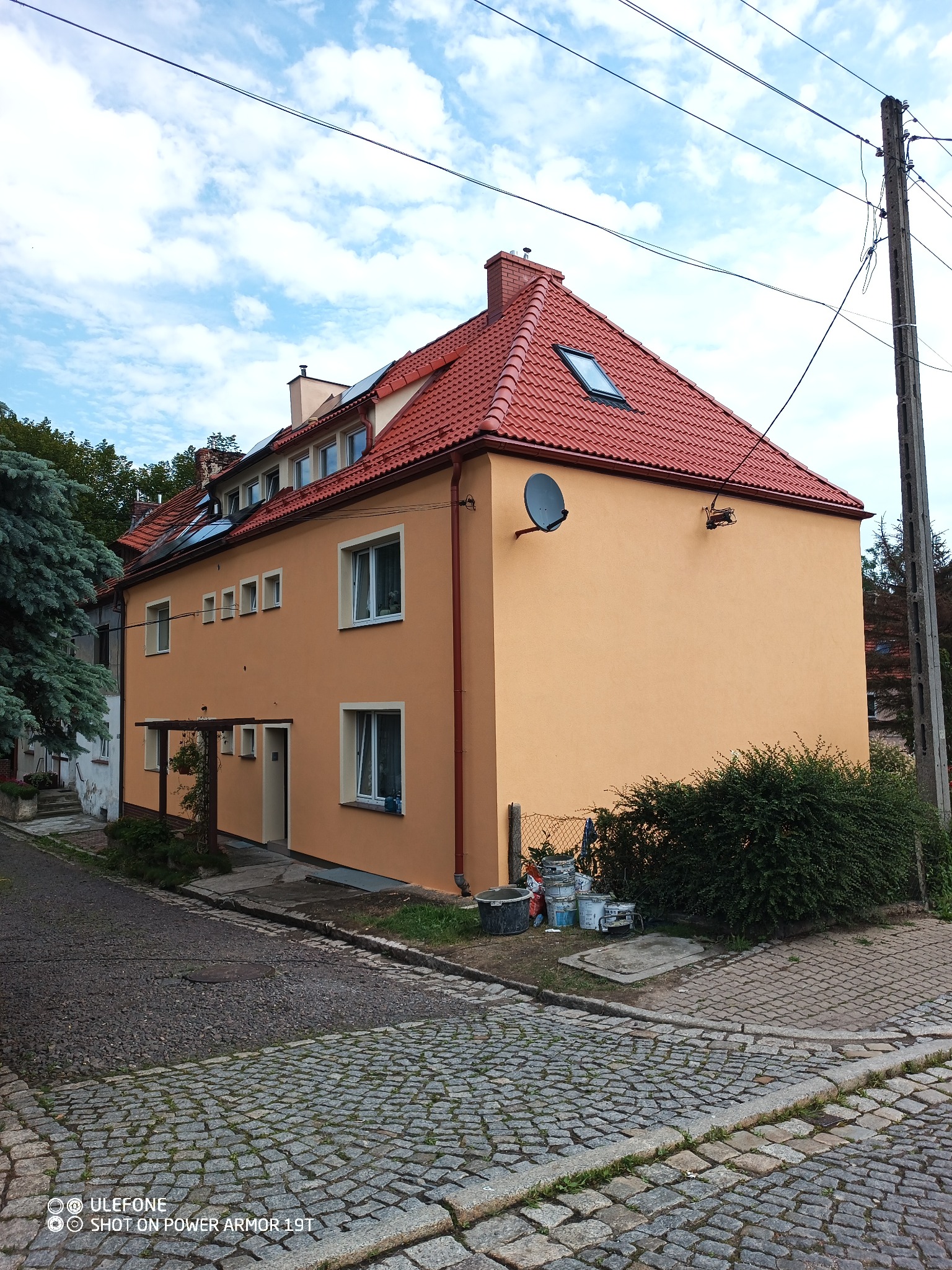 Dwupiętrowy budynek z pomarańczową elewacją i czerwoną dachówką, widoczny z poziomu brukowanej ulicy. W tle błękitne niebo z chmurami. Przy ścianie budynku widoczne wiaderka z farbą i krzew.