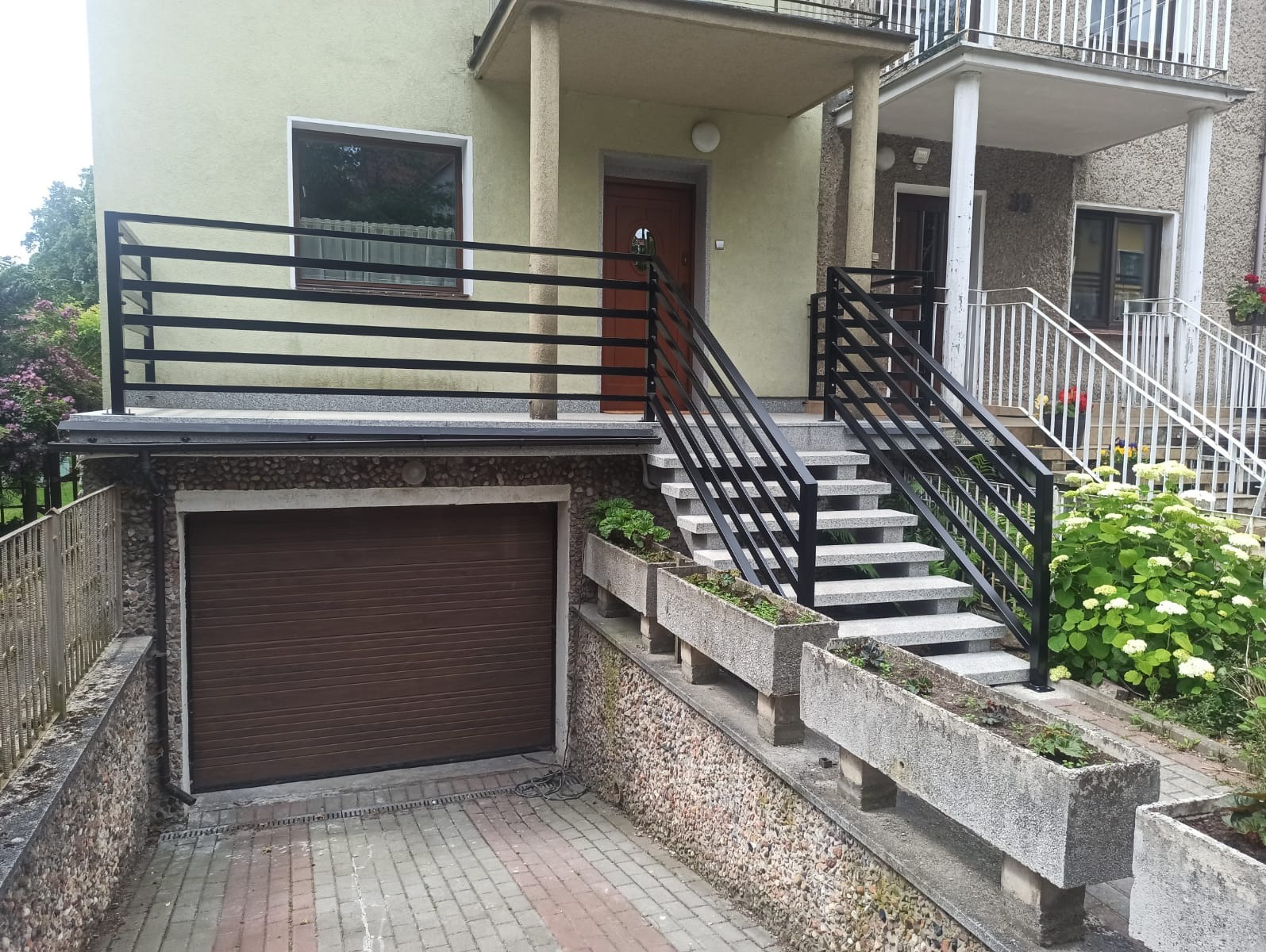 Dwukondygnacyjny dom z elewacją w odcieniach beżu i szarości, z brązową bramą garażową i czarnymi balustradami schodów wejściowych, betonowe donice wzdłuż schodów.