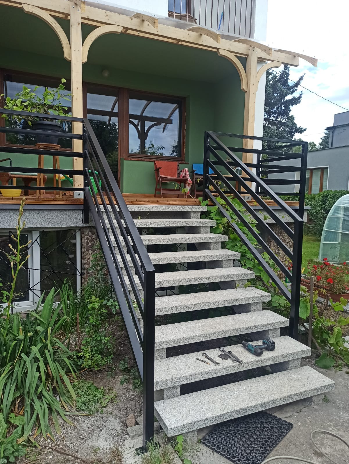 Schody zewnętrzne z szarego granitu z czarną metalową balustradą, narzędzia na stopniach, zielona elewacja domu w tle.