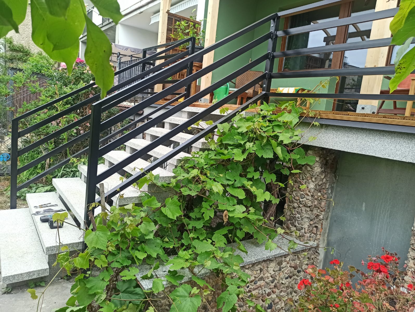 Betonowe schody zewnętrzne z czarną, metalową balustradą, częściowo porośnięte winoroślą, prowadzące do wejścia na podwyższony taras.