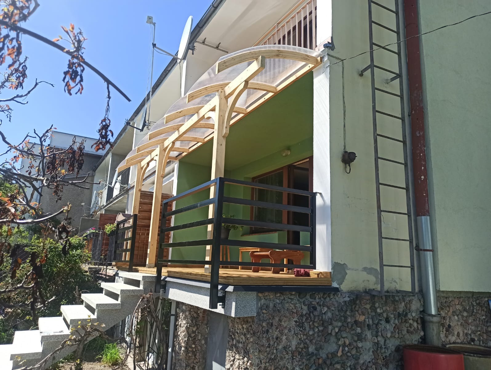 Zadaszony taras z drewnianą konstrukcją i czarną balustradą, widoczne schody wejściowe i drabina na ścianie budynku.