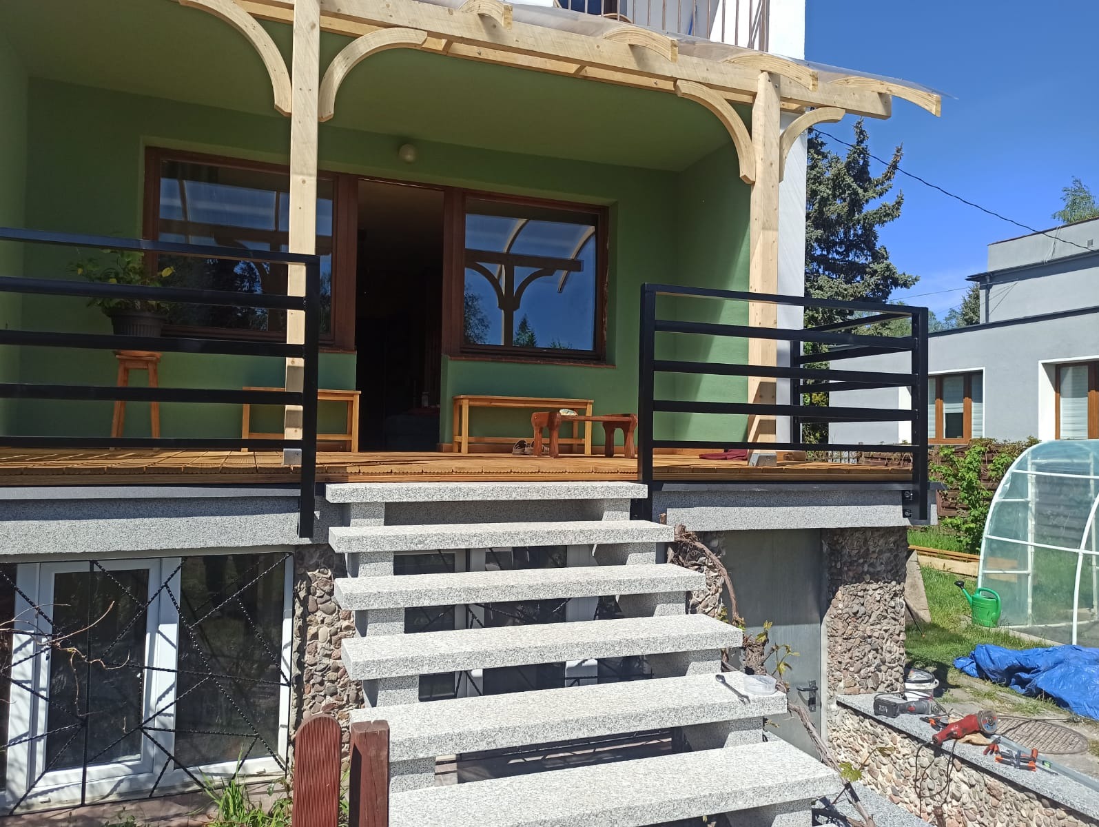 Zielony dom z drewnianą pergolą, granitowymi schodami i czarną balustradą, widok z dołu, narzędzia na ziemi sugerują trwające prace wykończeniowe.
