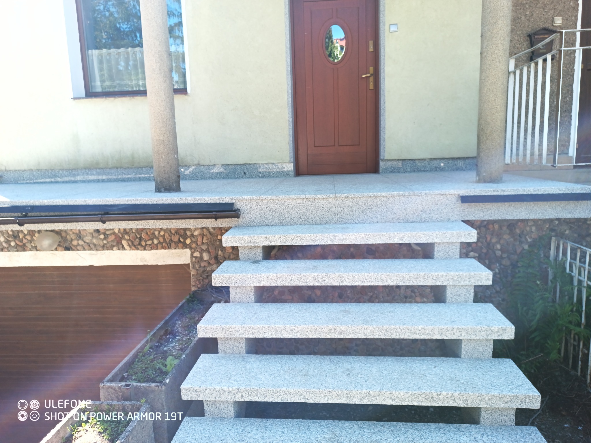Wejście do domu z granitowymi schodami, brązowe drzwi z owalnym okienkiem, kolumny po bokach, elewacja w odcieniach beżu, widoczny fragment garażu i roślinności.