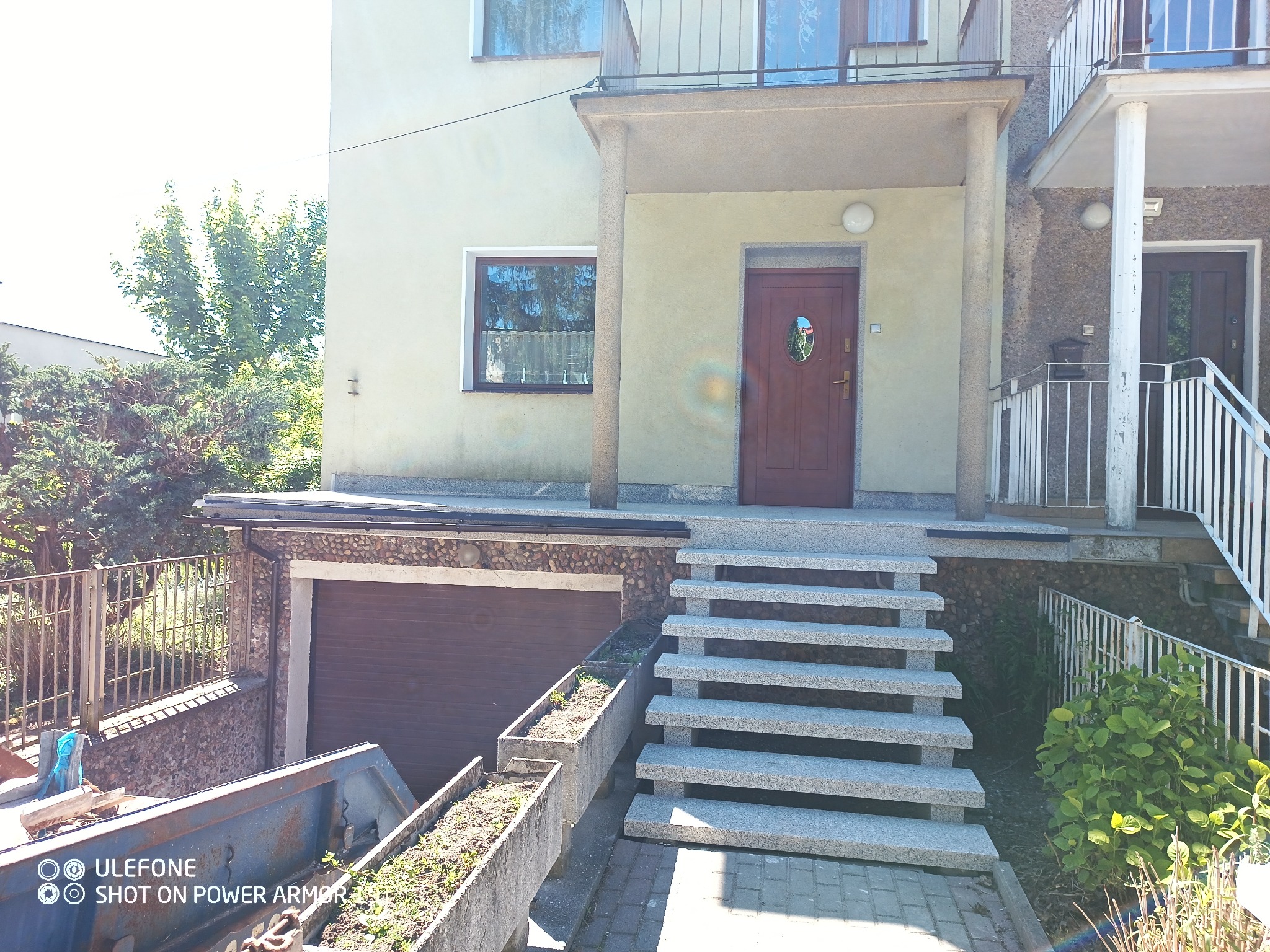 Dwukondygnacyjny dom z beżową elewacją, kamiennym wykończeniem garażu, betonowymi schodami wejściowymi i brązowymi drzwiami.