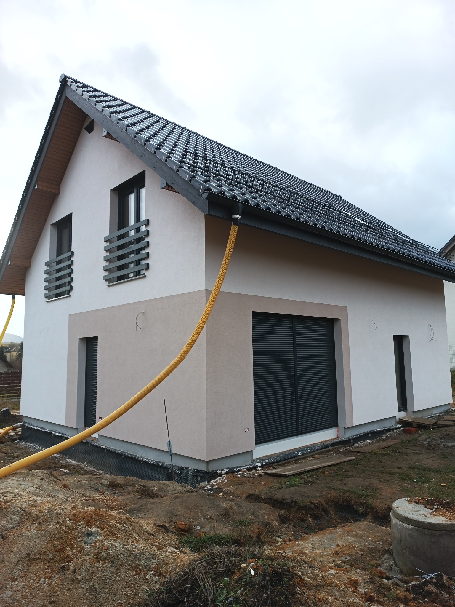 Nowo otynkowany dom jednorodzinny z ciemną dachówką i szarą stolarką okienną, widoczny w trakcie prac wykończeniowych terenu, z żółtym wężem roboczym przy elewacji.