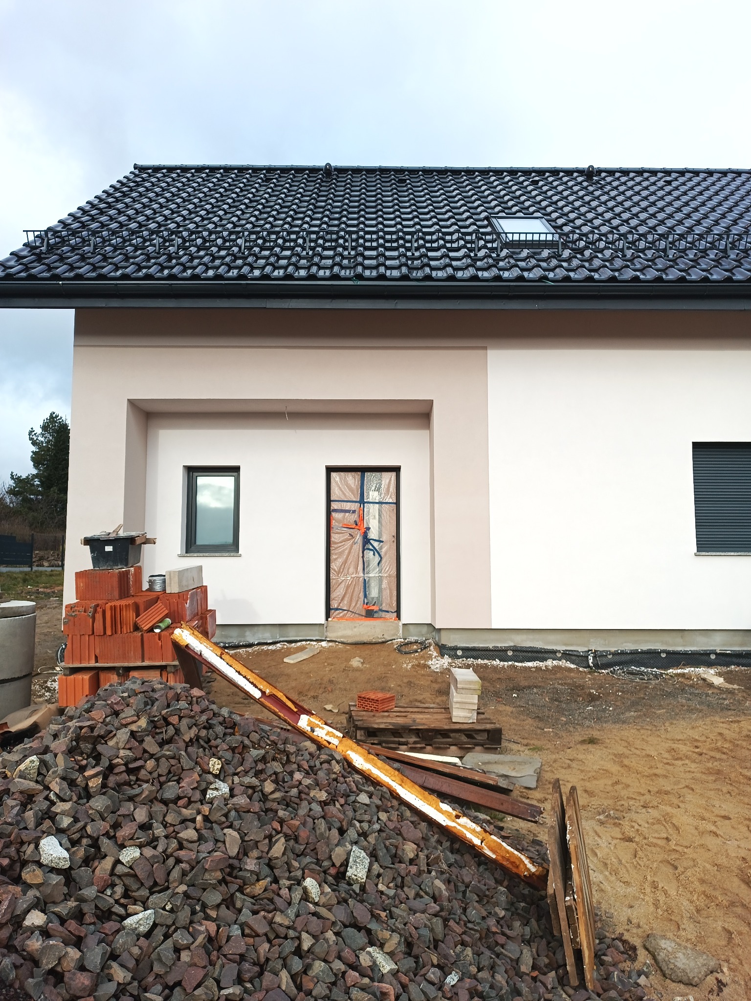 Nowo wybudowany dom jednorodzinny z jasną elewacją, czarną dachówką i zabezpieczonymi drzwiami wejściowymi, widoczne materiały budowlane i gruz.