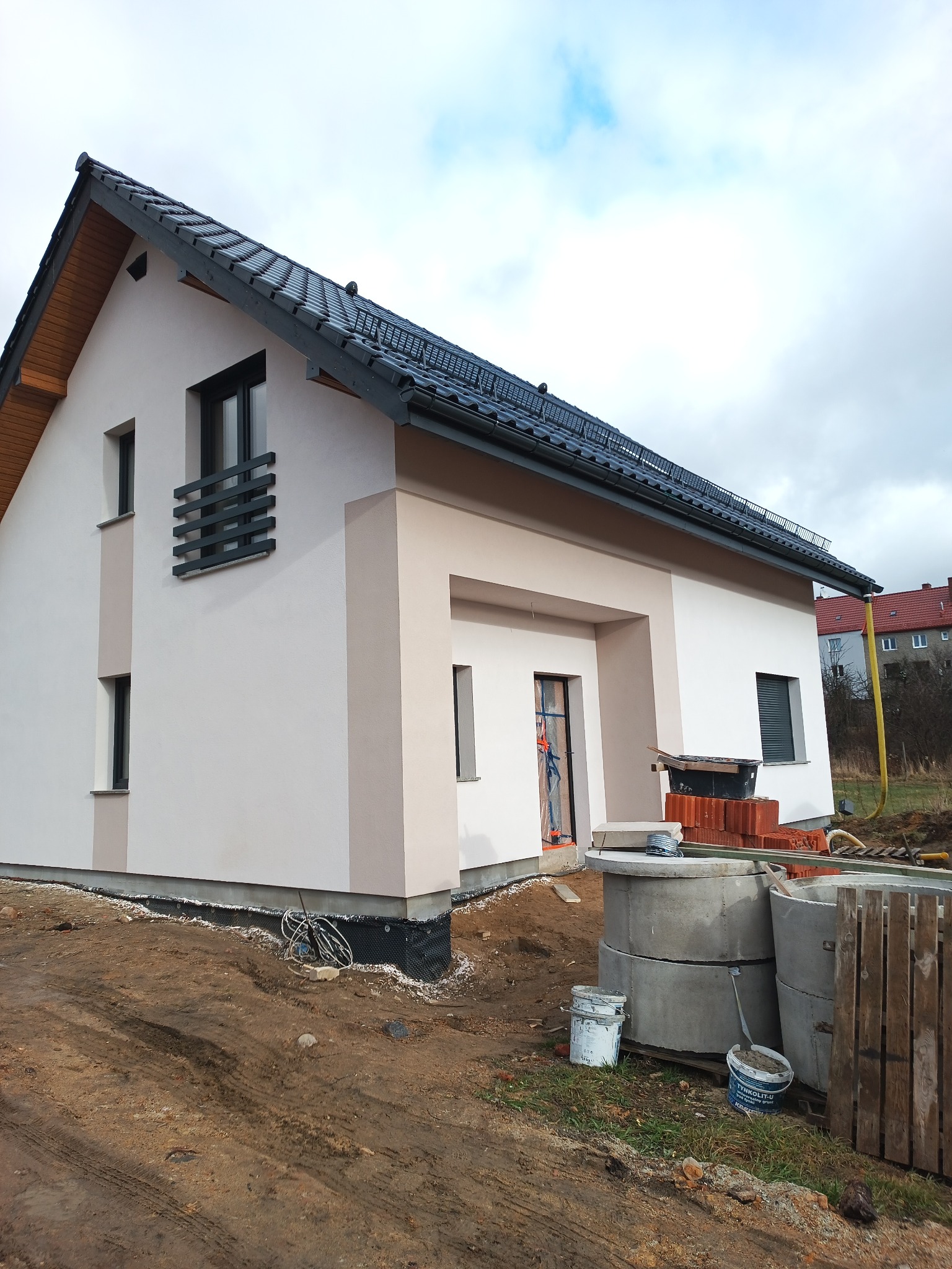 Nowoczesny dom jednorodzinny z elewacją w odcieniach beżu i bieli, czarny dach i okna, teren wokół domu w trakcie prac wykończeniowych, widoczne materiały budowlane i narzędzia.