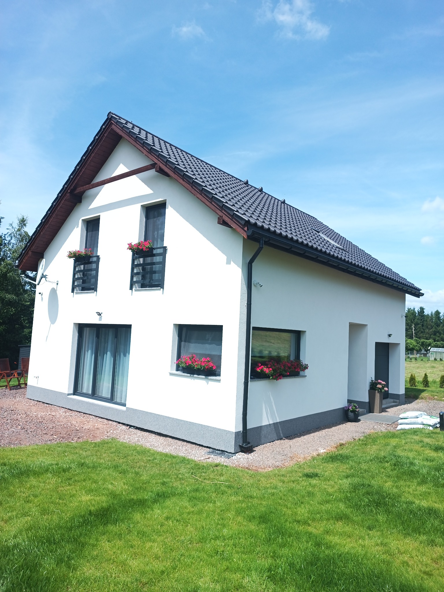 Biały dom z ciemną dachówką i szarą podmurówką, ozdobiony kwiatami w oknach i na balkonach. Widok na elewację frontową w słoneczny dzień.