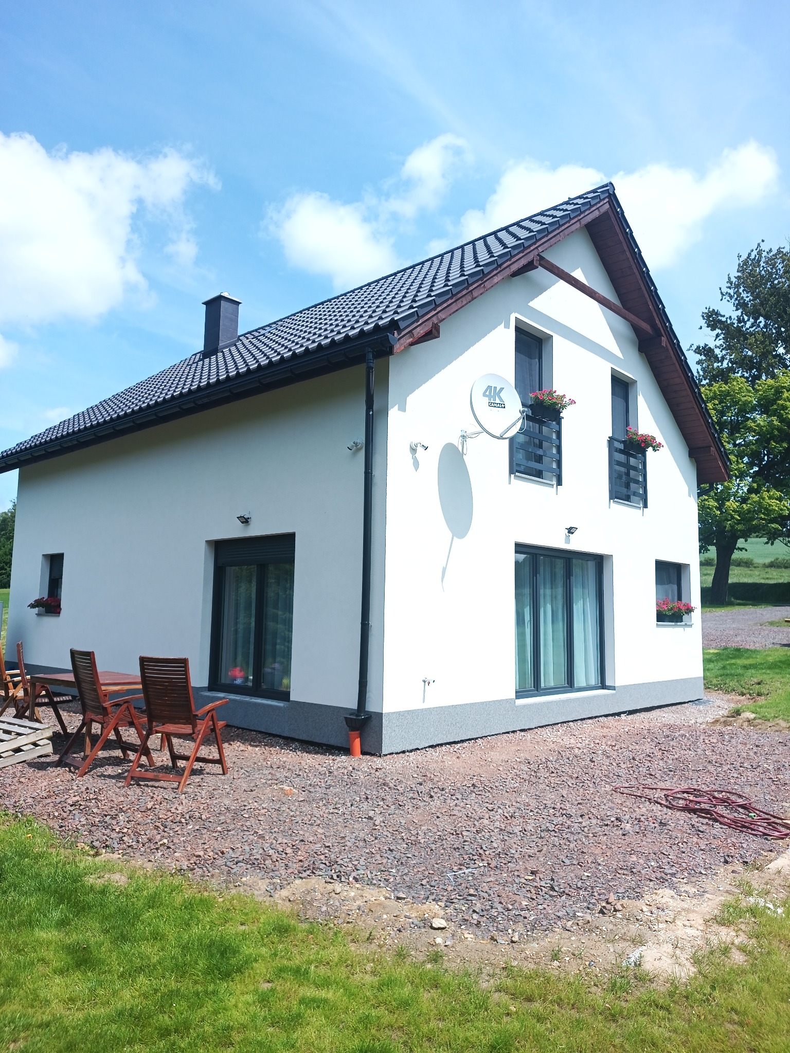 Biały dom z szarym dachem i czarnymi oknami, otoczony zielenią i kamieniami. Na ścianie satelita 4K. Widoczne drewniane meble ogrodowe.