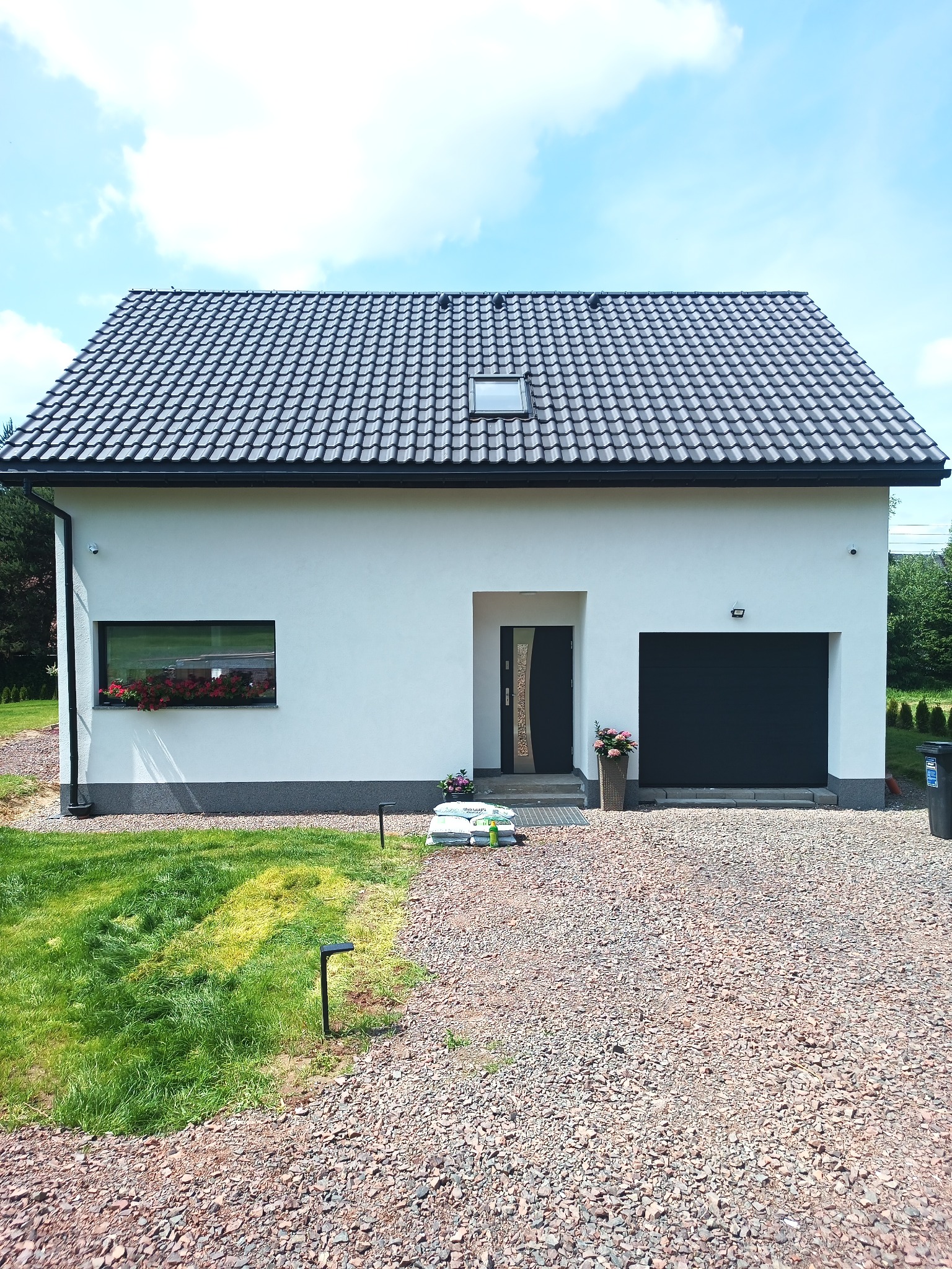 Biały dom z ciemną dachówką i garażem, okno z kwiatami, podjazd z kamienia. Widoczne worki z materiałami budowlanymi i elementy oświetlenia ogrodu.