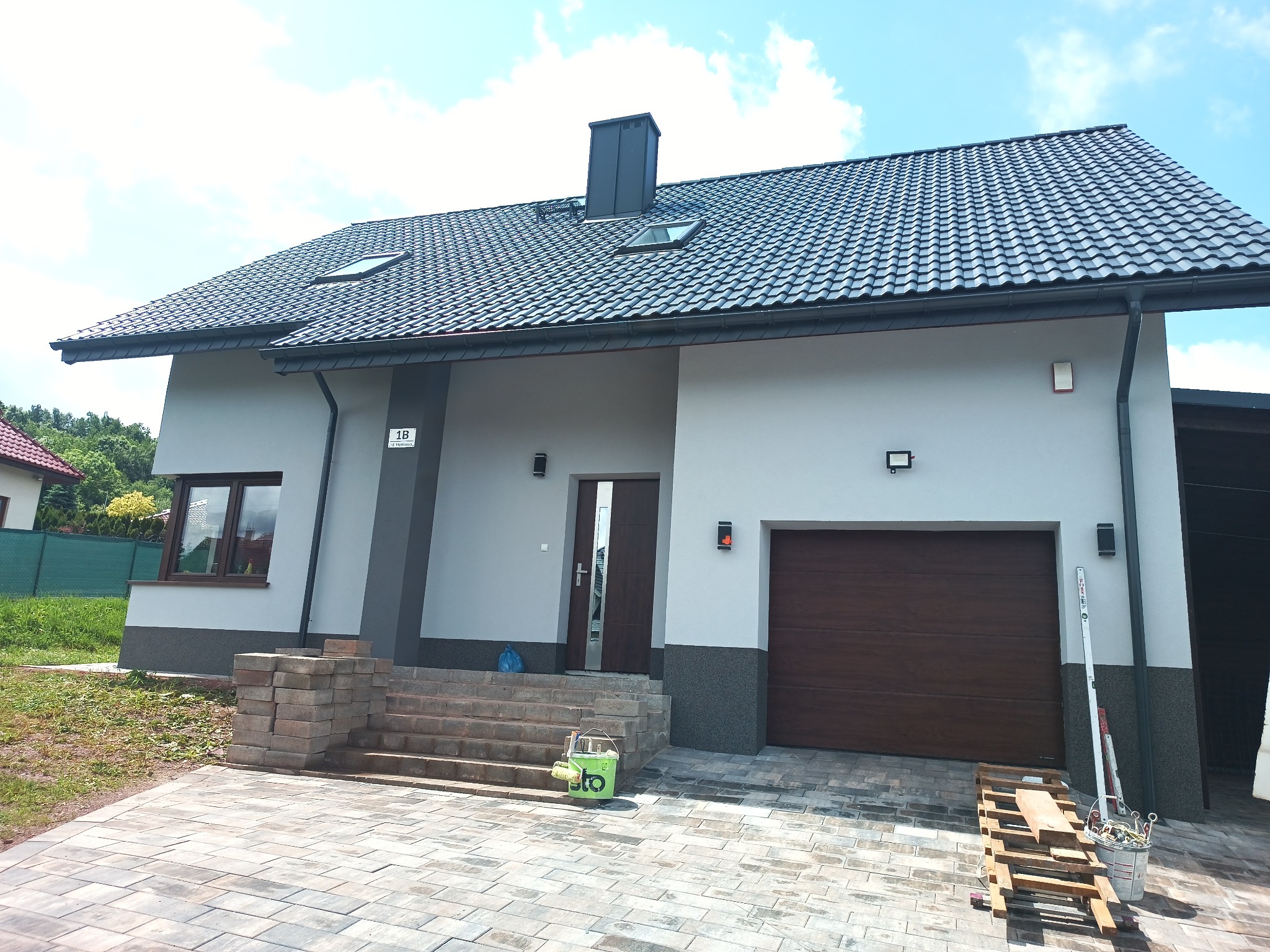 Nowoczesny dom jednorodzinny z szarą elewacją, grafitowym dachem i brązową bramą garażową. Widoczne narzędzia malarskie sugerują prace wykończeniowe.