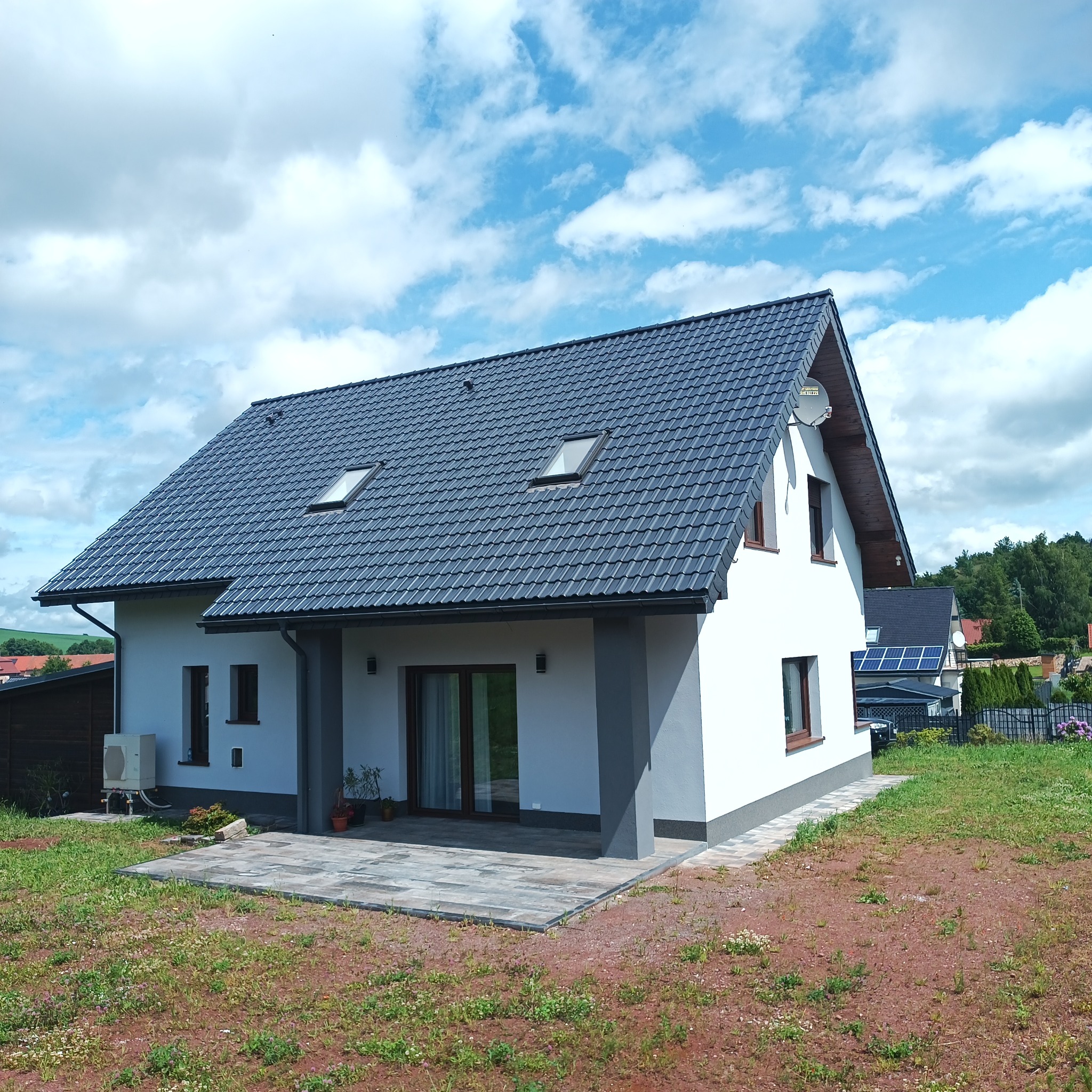 Nowoczesny, parterowy dom z ciemną dachówką, białymi ścianami i szarymi akcentami. Widoczny taras z kostki brukowej i zadbany trawnik. W tle błękitne niebo z chmurami.