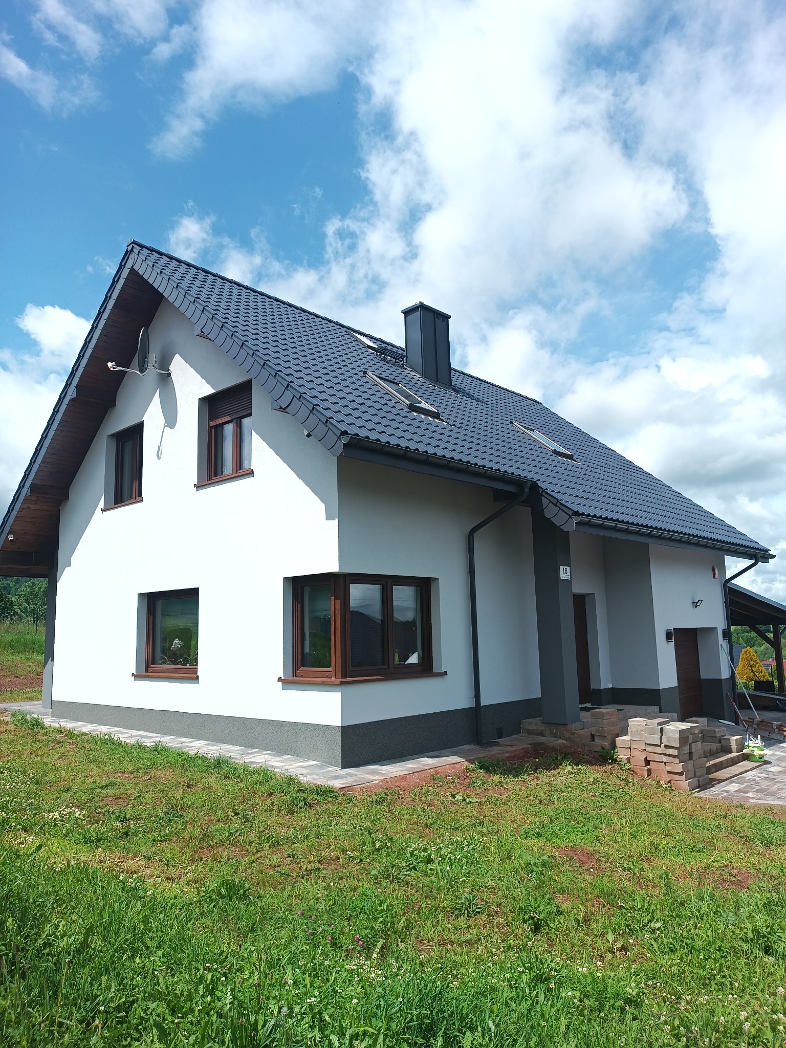Biały dom z ciemną dachówką i brązowymi oknami na tle błękitnego nieba z chmurami. Świeżo położona kostka brukowa i trawnik. Widoczna antena satelitarna.