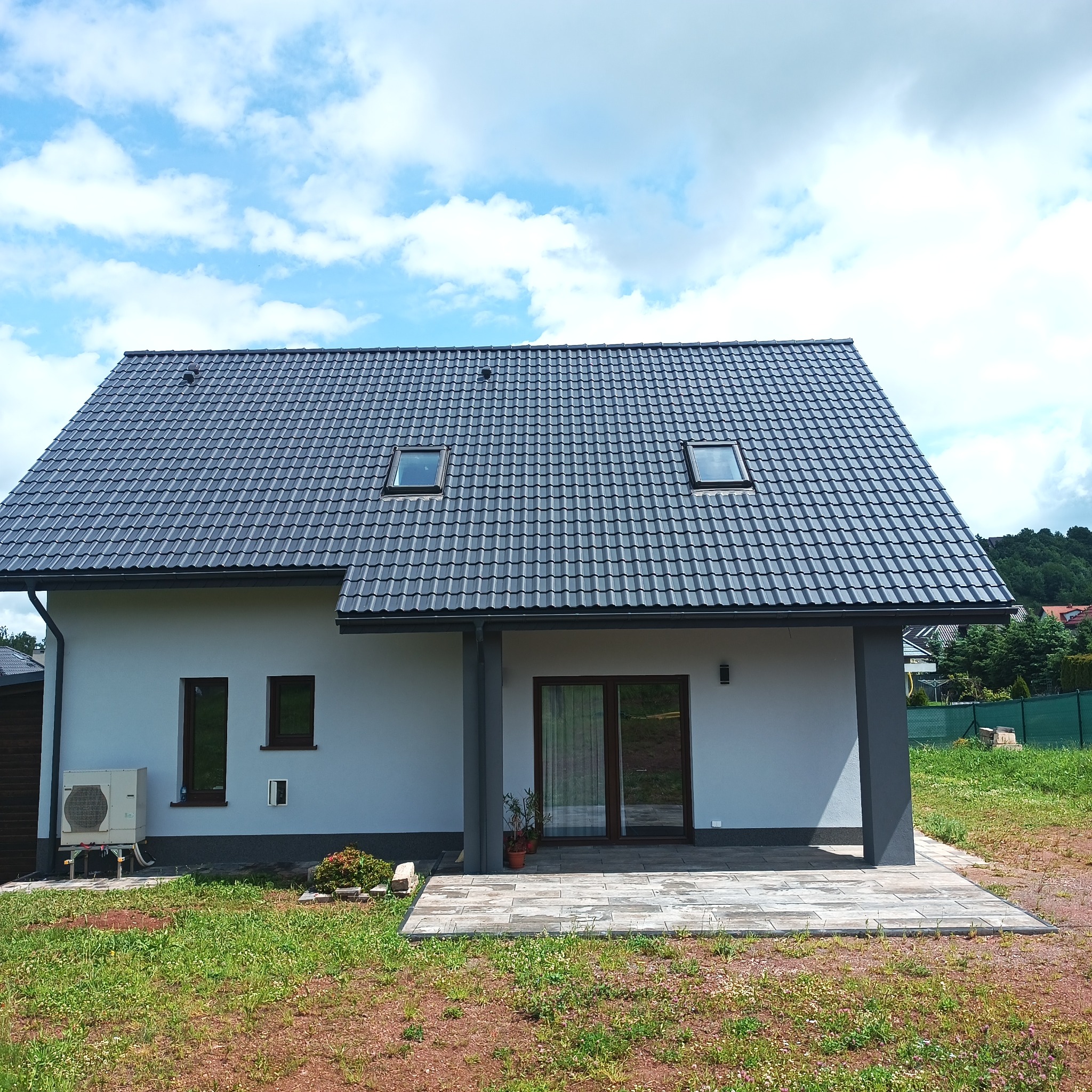 Nowoczesny dom jednorodzinny z szarą elewacją i grafitowym dachem z dachówką. Widoczne okna dachowe, taras z kostki brukowej i klimatyzator na zewnątrz budynku.